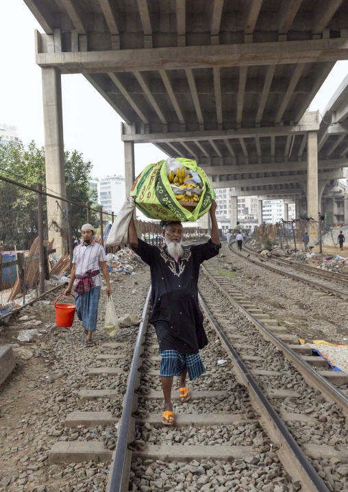 A bangladeshi old man walks along the rail track with a bag on his head, Dhaka Division, Dhaka, Bangladesh