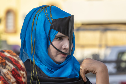 Close-up portrait of a young Omani woman wearing a bright blue headscarf with gold cord trim and a black leather face mask, Ad Dakhiliyah Region, Nizwa, Oman