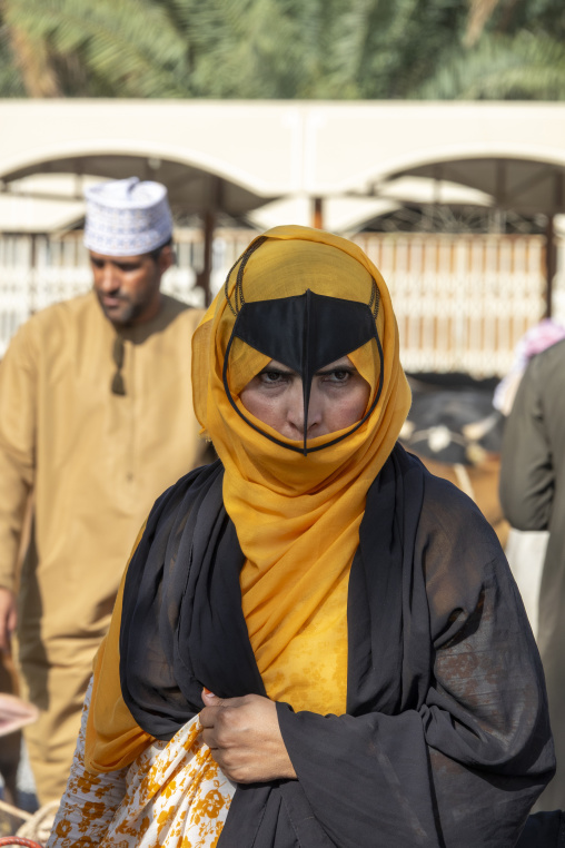 Close-up profile portrait of an Omani woman wearing a golden headscarf and black leather face mask against a dark blurred background, Ad Dakhiliyah Region, Nizwa, Oman