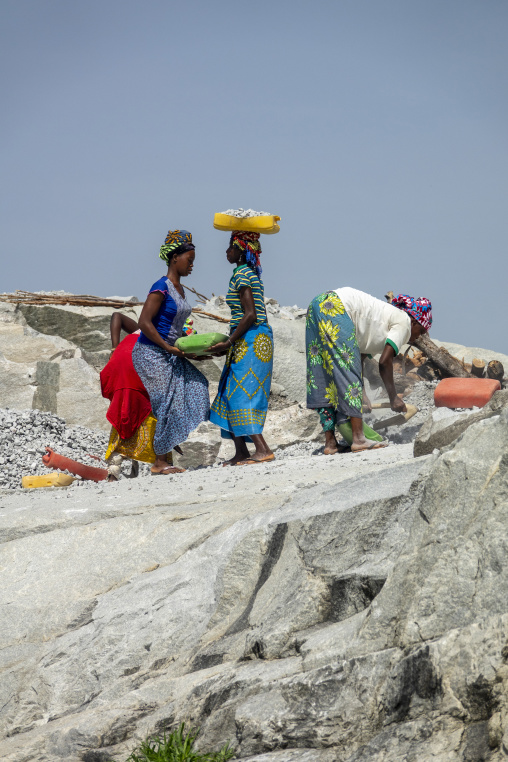 Women carry basins of crushed granite while a man digs with a shovel at the quarry, Savanes district, Shienlow, Ivory Coast