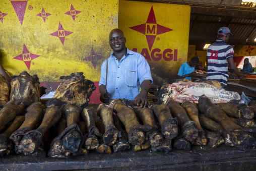 A smiling butcher stands behind rows of smoked animal legs and heads at an indoor market, Poro region, Korhogo, Ivory Coast