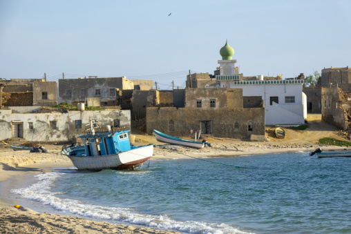 Weathered fishing boats beached in front of a traditional mudbrick village with a green-domed mosque minaret, Dhofar Governorate, Mirbat, Oman