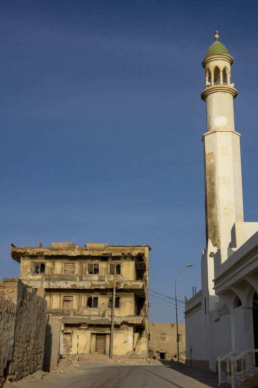 Ruined multi-story building standing beside a white mosque minaret with a green dome under a clear blue sky, Dhofar Governorate, Mirbat, Oman