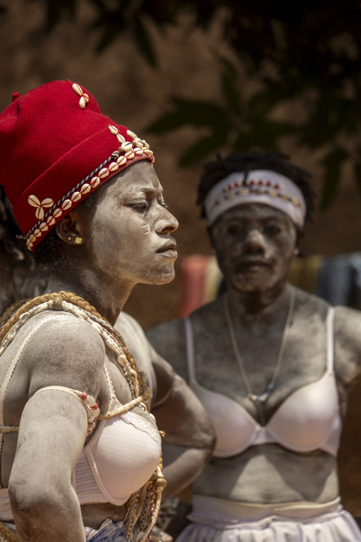 A woman in a red cowrie-shell hat stands before another during the Adjoua Messouma Komians initiation ceremony, Moyen-Comoé, Aniassue, Ivory Coast