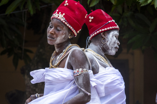 Two performers stand back to back wearing red cowrie-shell hats during the Adjoua Messouma Komians initiation ceremony, Moyen-Comoé, Aniassue, Ivory Coast
