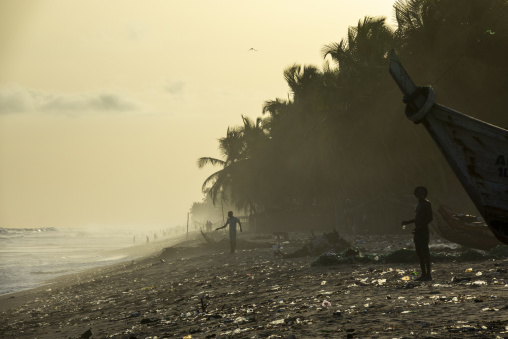 Fishermen stand on a littered beach beside a boat in N'zima village, Sud-Comoé, Grand-Bassam, Ivory Coast
