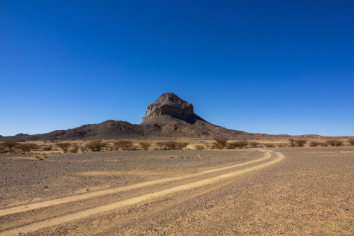 Dirt road winding toward a rocky butte in the Sahara desert, North Africa, Tamanrasset, Algeria