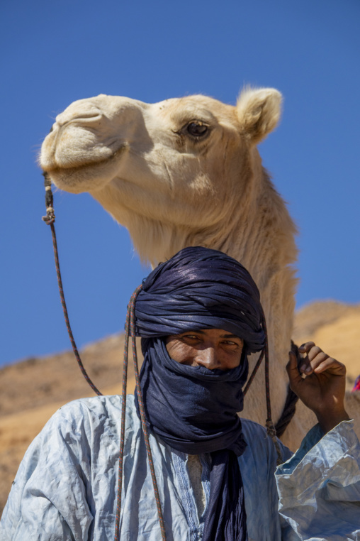 Tuareg man in an indigo turban standing beneath his camel's head in the desert, North Africa, Tamanrasset, Algeria
