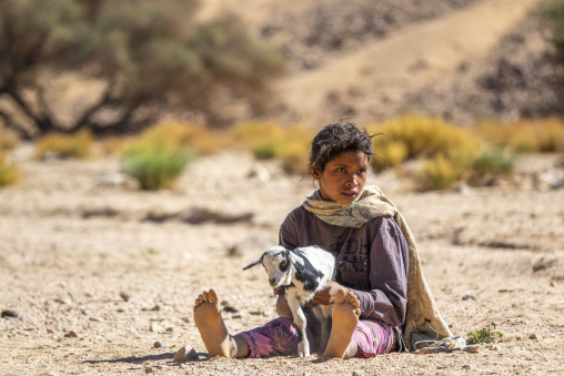 Young girl sitting on the ground holding a baby goat in the desert, North Africa, Tamanrasset, Algeria