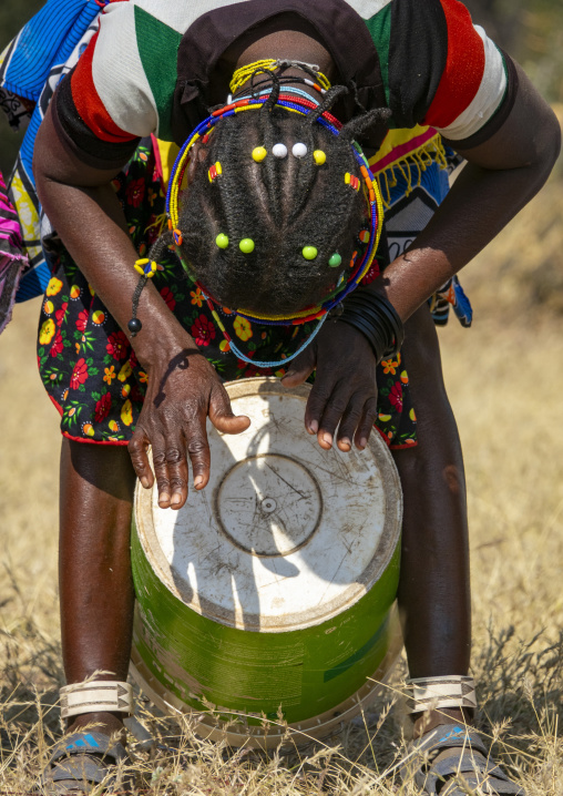 Mudimba tribe woman bending forward to play a drum made from a repurposed metal barrel while wearing a beaded headdress, Cunene Province, Cahama, Angola
