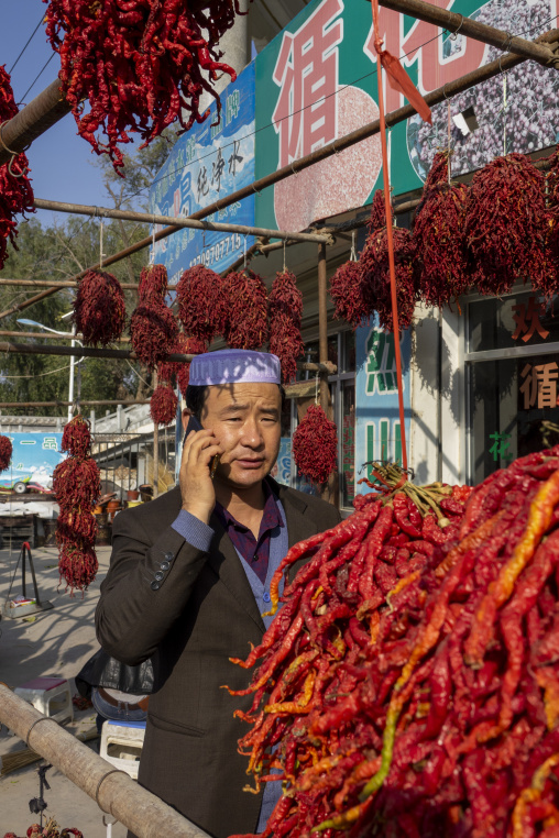 A man wearing a prayer cap talks on his phone beside hanging red chili peppers, Tongren County, Rebkong, China