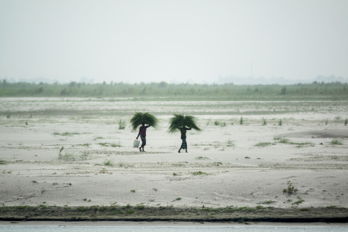 Two men carrying bundles of grass on their heads across a river