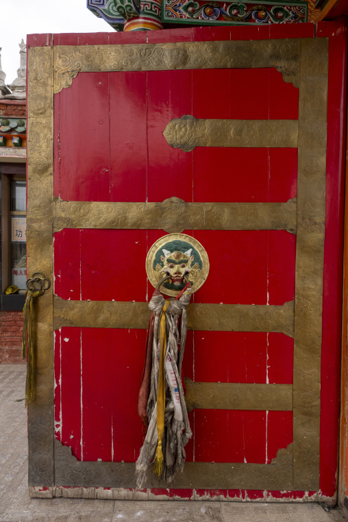 An ornate red door with a gilded guardian mask and prayer scarves stands at Hezuo monastery, Gansu Province, Hezuo, China