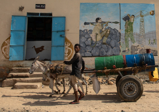 Eritrean boy carrying water on a cart in front of a mural depciting the war, Gash-Barka, Hagaz, Eritrea
