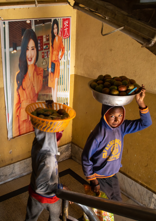 Eritrean boys carrying fruits on their heads in a stair decorated with a chinese poster, Central region, Asmara, Eritrea