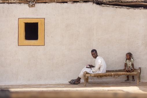 A man uses his phone while sitting on a rope bed beside a young girl in a courtyard with a painted yellow window, Red Sea State, Port Sudan, Sudan