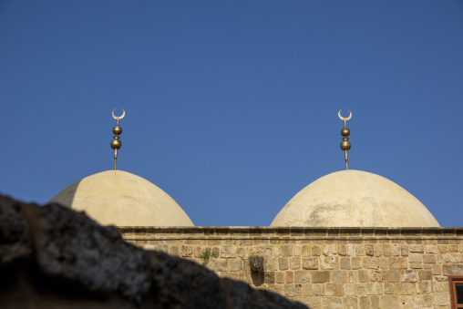 Twin mosque domes topped with golden crescent finials rise above an old stone wall against a clear sky, South Governorate, Tyre, Lebanon