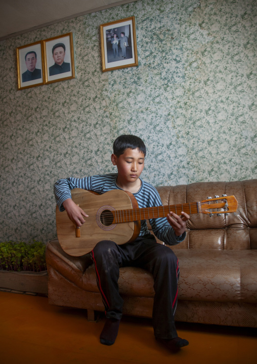 Boy playing guitar in front of the portraits of the Leaders, North Hamgyong, Jung Pyong Ri, North Korea