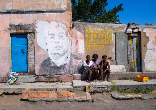 Angolan women sit in front of an old propaganda wall painting with Leonid Brejnev, Cunene Province, Cahama, Angola