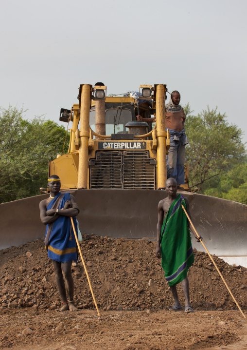 Two Indigenous Mursi Men Standing In Front Of A Caterpillar Steam Shovel On Coated Road  Construction Work In Mago Park Omo Valley  Ethiopia