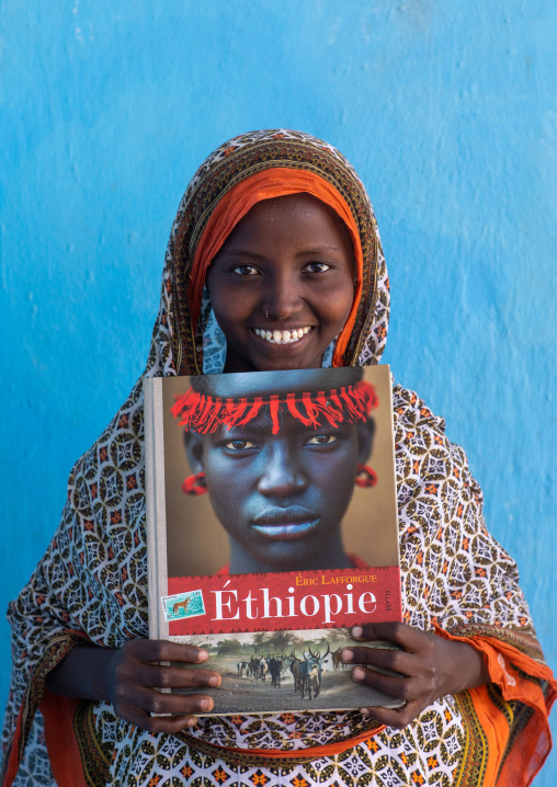 Portrait of a veiled smiling afar tribe girl with sharpened teeth holding eric lafforgue book, Afar Region, Afambo, Ethiopia