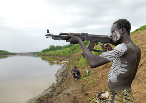 Mid Adult White Painted Chest Karo Man Aiming With Kalashnikov Rifle Near Omo River Back Of Hamer Shepherd Ethiopia