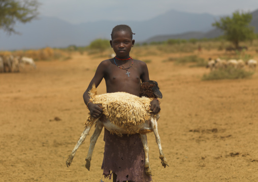 Tsemay tribe teenage girl carrying a sheep in her arms in a desert landscape, Omo valley, Ethiopia