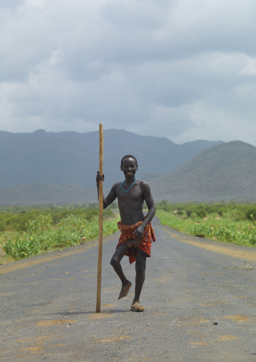 Disabled tsemay tribe boy walking on coated road with a pole in his hand and smiling, Omo valley, Ethiopia