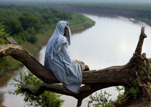 Karo Tribe Man In The Morning Sitting On A Tree Trunk Over The Omo River, Korcho Village, Omo Valley, Ethiopia