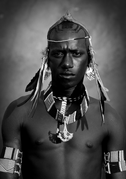 Black and white portrait of a young tsamay whipper with fierce look, Feathers attached to his hairband and traditional necklace, Turmi, Ethiopia