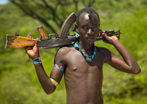 Young tsemay tribe man with beaded jewels carrying kalashnikov rifle on his shoulders, Omo valley, Ethiopia