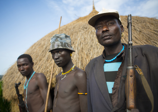 Menit tribe men displaced by governement holding guns in their arms in front of their new houses, Jemu, Ethiopia