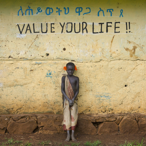 Young shy Suri boy leaning against the wall of a school covered with amharic and english writing, Tulgit, Omo valley, Ethiopia