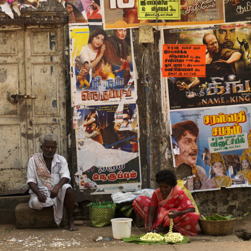 Man Sitting Next To His Wife Making Flowers Garland In Front Of Bollywood Posters, Madurai, India