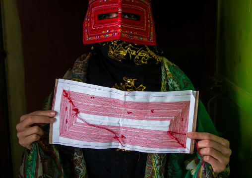 a bandari woman wearing a traditional mask called the burqa and showing one she is sewing, Hormozgan, Minab, Iran