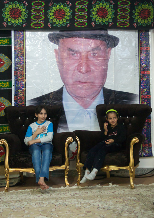 Girls sit on chairs in front of the giant portrait of a member of their family who died during Muharram, Lorestan Province, Khorramabad, Iran