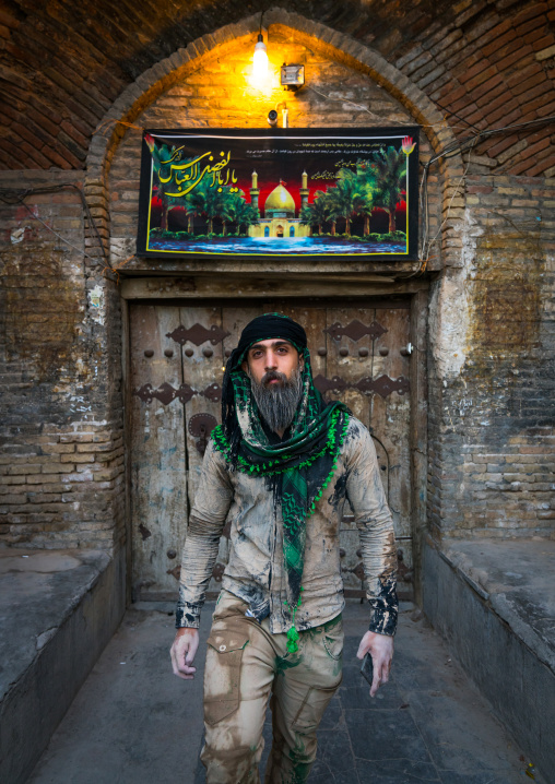 Iranian shiite muslim man standing in front of an old wooden door after rubbing mud on his clothes during the Kharrah Mali ritual to mark the Ashura ceremony, Lorestan Province, Khorramabad, 