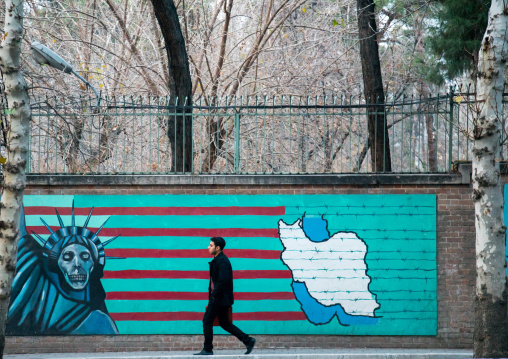 man passing in front of anti-american mural propoganda slogan depict statue liberty skeleton on the wall of the former united states embassy, Central district, Tehran, Iran