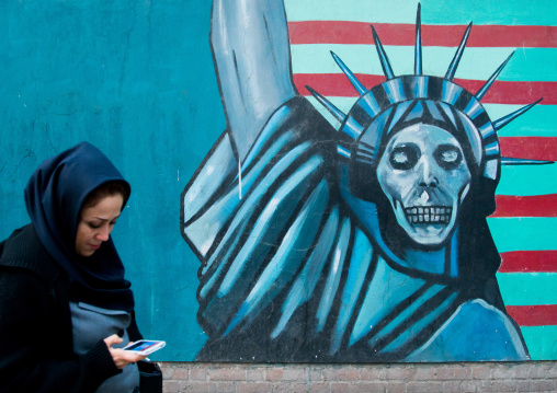 woman with phone passing in front of an anti-american propoganda slogan depicting statue liberty skeleton on the wall of the united states embassy, Central district, Tehran, Iran