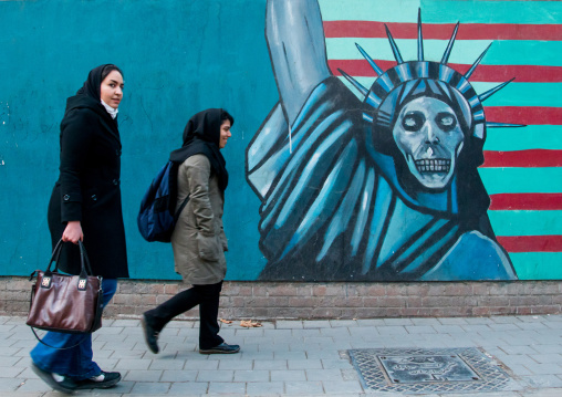 women passing in front of anti-american mural propoganda slogan depict statue liberty skeleton on the wall of the former united states embassy, Central district, Tehran, Iran