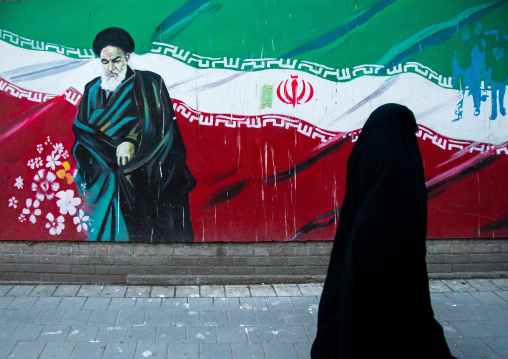 veiled woman passing in front of propaganda sign with ayatollah khomeini on the wall of the former u.s. embassy, Central district, Tehran, Iran
