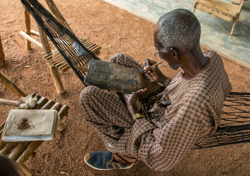 Senior man sit on a hammock writing koran on a wood board, Savanes district, Kouto, Ivory Coast
