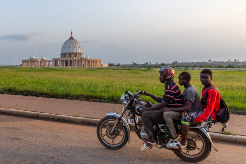 African men on a motorbike passing in front of our lady of peace basilica christian cathedral, Région des Lacs, Yamoussoukro, Ivory Coast