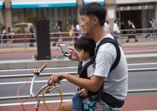 Father looking at his smart phone while riding a bicycle with his child, Kanto region, Tokyo, Japan
