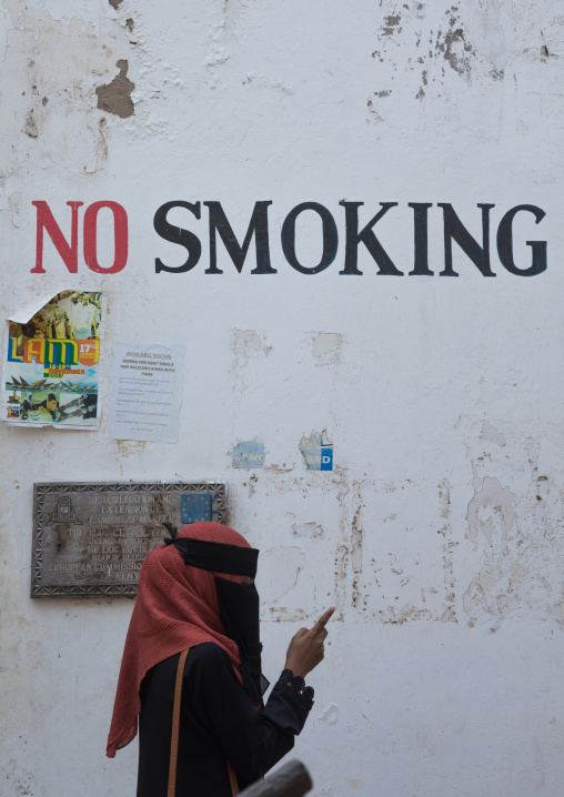 Muslim woman in burqa under a no smoking sign on a wall using her mobile phone, Lamu county, Lamu town, Kenya