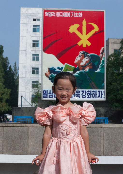 North Korean girl in front of a propaganda billboard in the street with the workers' Party of North Korea logo, Pyongan Province, Pyongyang, North Korea