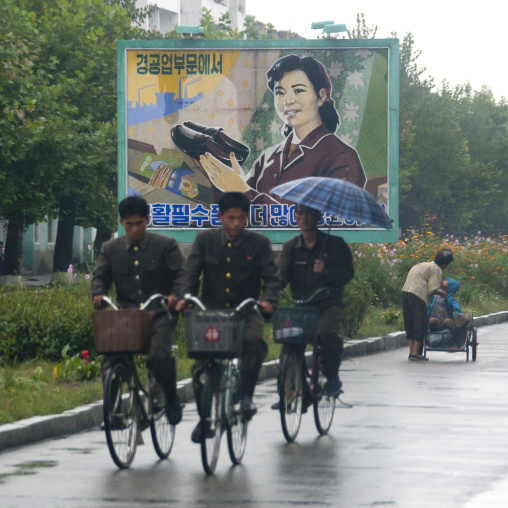 North Korean soldiers riding bicycles on a wet road with an advertising poster for shoes in background, South Hamgyong Province, Hamhung, North Korea