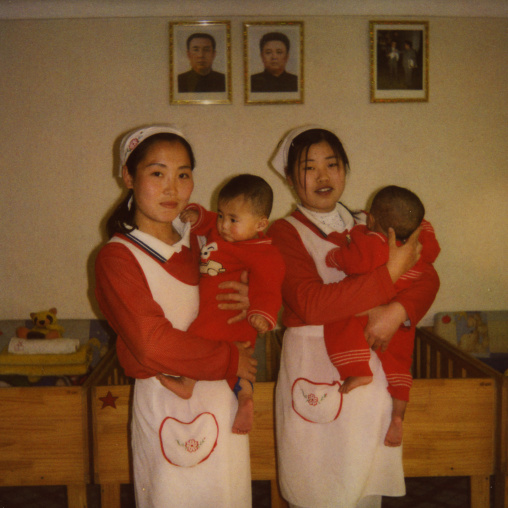 Polaroid of nurses taking care of babies in an orphanage under the official portraits of the Dear Leaders, South Pyongan Province, Nampo, North Korea