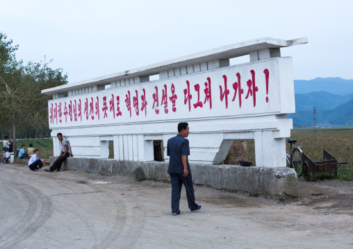 North Korean man passing in front of a propaganda billboard saying let's push revolution and construction as the spirit of the great leader in his lifetime, South Hamgyong Province, Hamhung, 