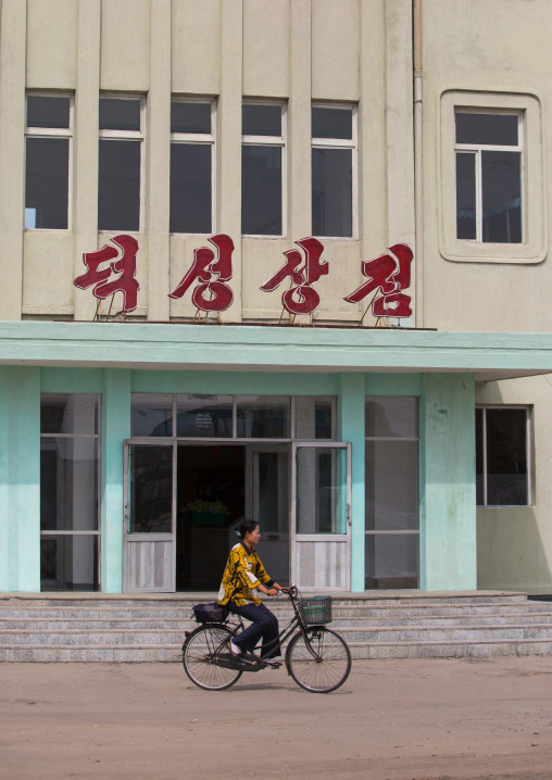 North Korean woman riding a bicycle in front of deoksong shop, South Hamgyong Province, Hamhung, North Korea